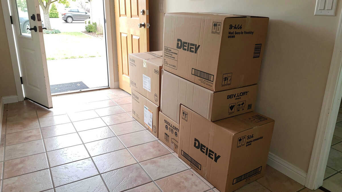 A photograph of a stack of cardboard delivery boxes on a tiled hallway floor beside a front door, daylight coming through the doorway