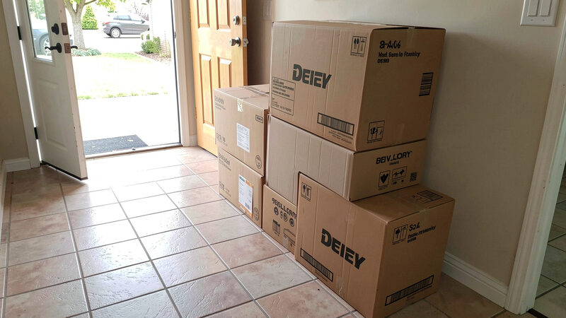 A photograph of a stack of cardboard delivery boxes on a tiled hallway floor beside a front door, daylight coming through the doorway
